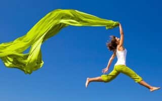 A person in green pants and a white top jumps in the air, holding a flowing green fabric against a clear blue sky.