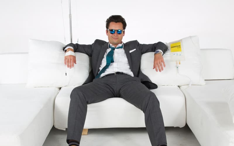 A man in a suit and sunglasses sits casually on a white sofa with arms spread, surrounded by white cushions.