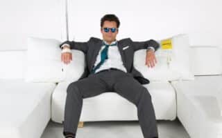 A man in a suit and sunglasses sits casually on a white sofa with arms spread, surrounded by white cushions. A man in a suit and sunglasses sits casually on a white sofa with arms spread, surrounded by white cushions.