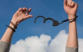 A pair of hands raised against a blue sky with clouds; one wrist is shackled to open handcuffs. A pair of hands raised against a blue sky with clouds; one wrist is shackled to open handcuffs.