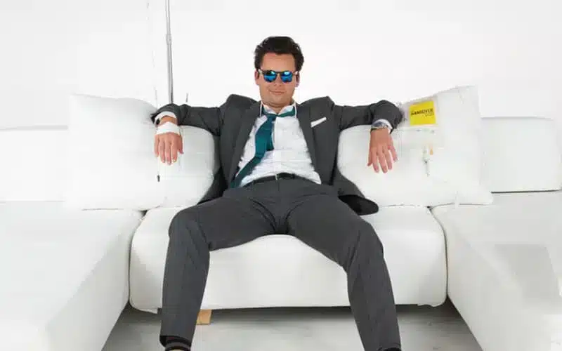 A man in a suit and sunglasses sits casually on a white sofa with arms spread, surrounded by white cushions.